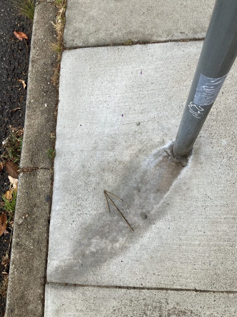 Stick in shape of arrow pointing up and to the left, on the sidewalk next to a pole. 