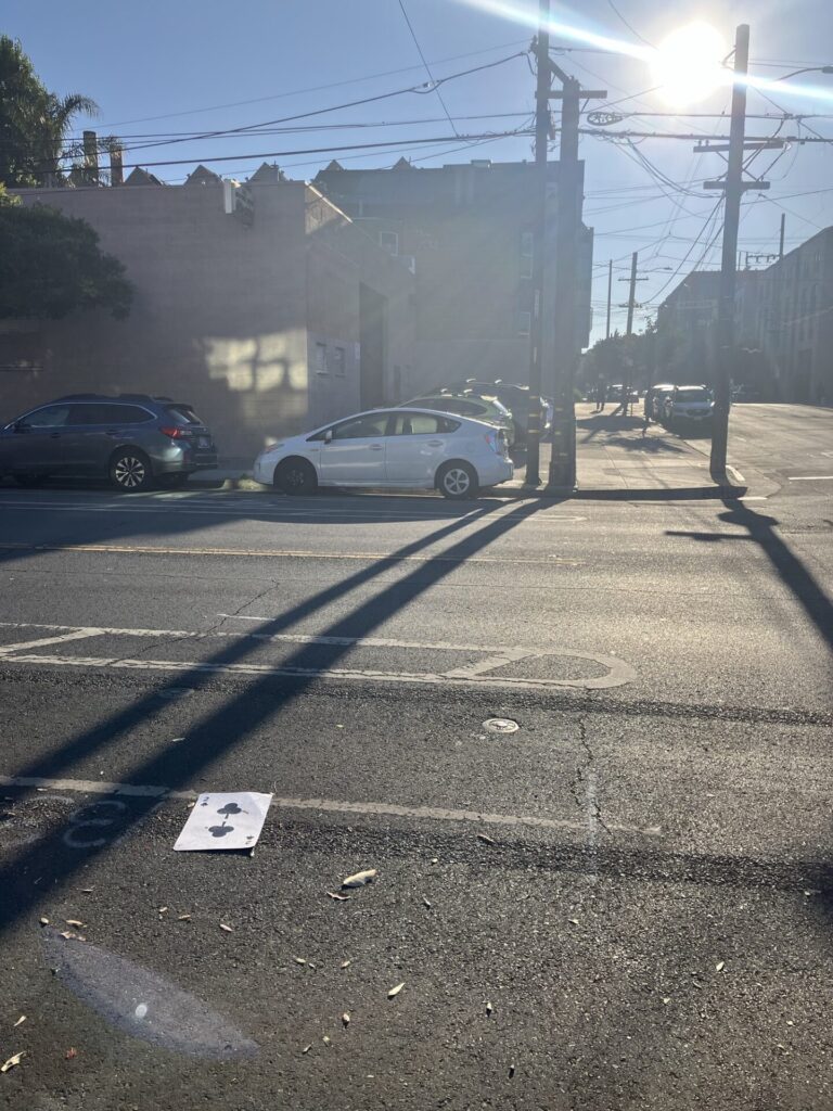 Sunny street with buildings, cars, and electrical poles in the background. In the street, a giant playing card, two of clubs. 